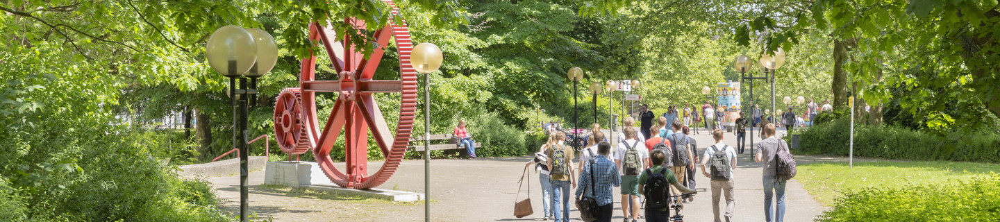 Studierende laufen über den Campus. Es ist eine große rote Skulptur in Form von großen Zahnrädern zu erkennen.