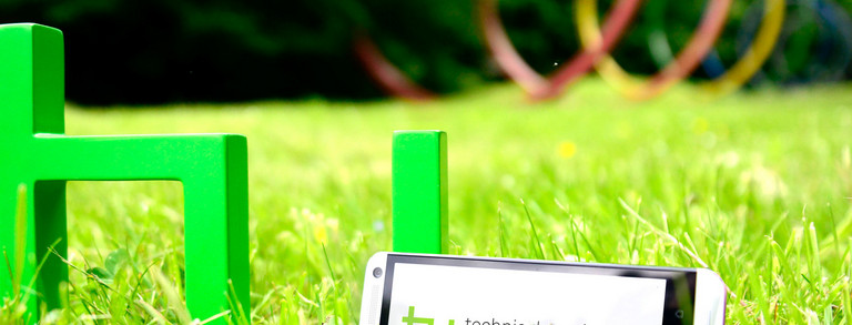 The green logo of TU Dortmund University is standing on a meadow. A smartphone is leaning against it.